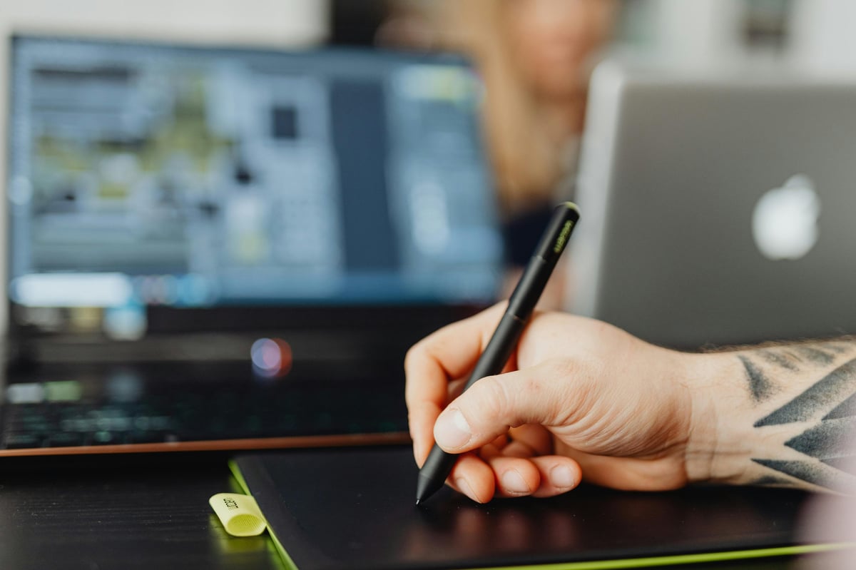 Person with tattooed arm holding a stylus pen over a graphics tablet with computer monitors and laptop in background